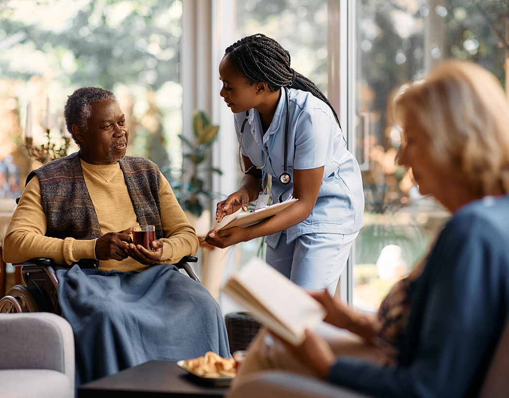 Black senior man in wheelchair drinking tea while talking to a nurse at residential care home.